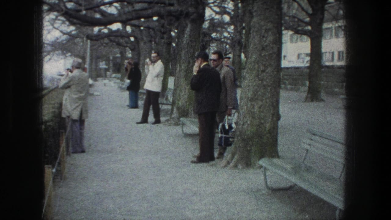 Adult men stand on a tree-lined path with benches and a stone wall nearby.