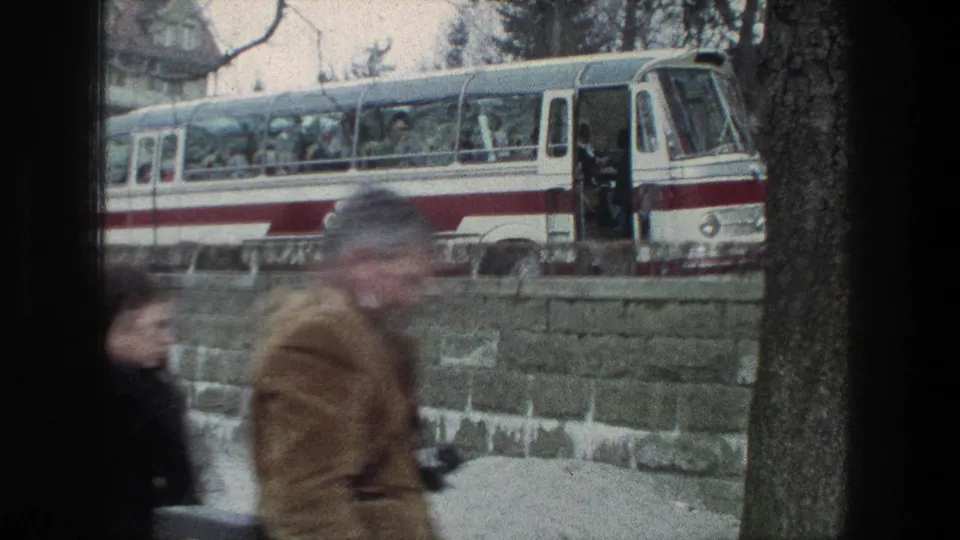 A red-and-white coach bus with passengers or boarding figures appears beyond a stone wall in Zurich, while blurred pedestrians pass in the foreground.