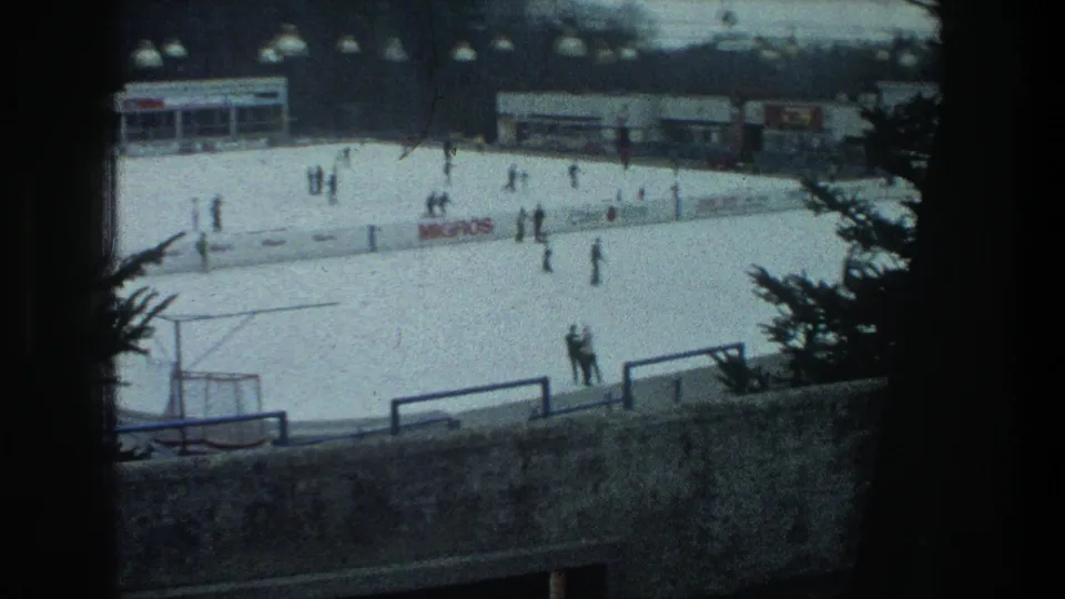 Elevated 1976 Zurich view of an outdoor ice rink with skaters, rink boards, trees, and low buildings in the background.