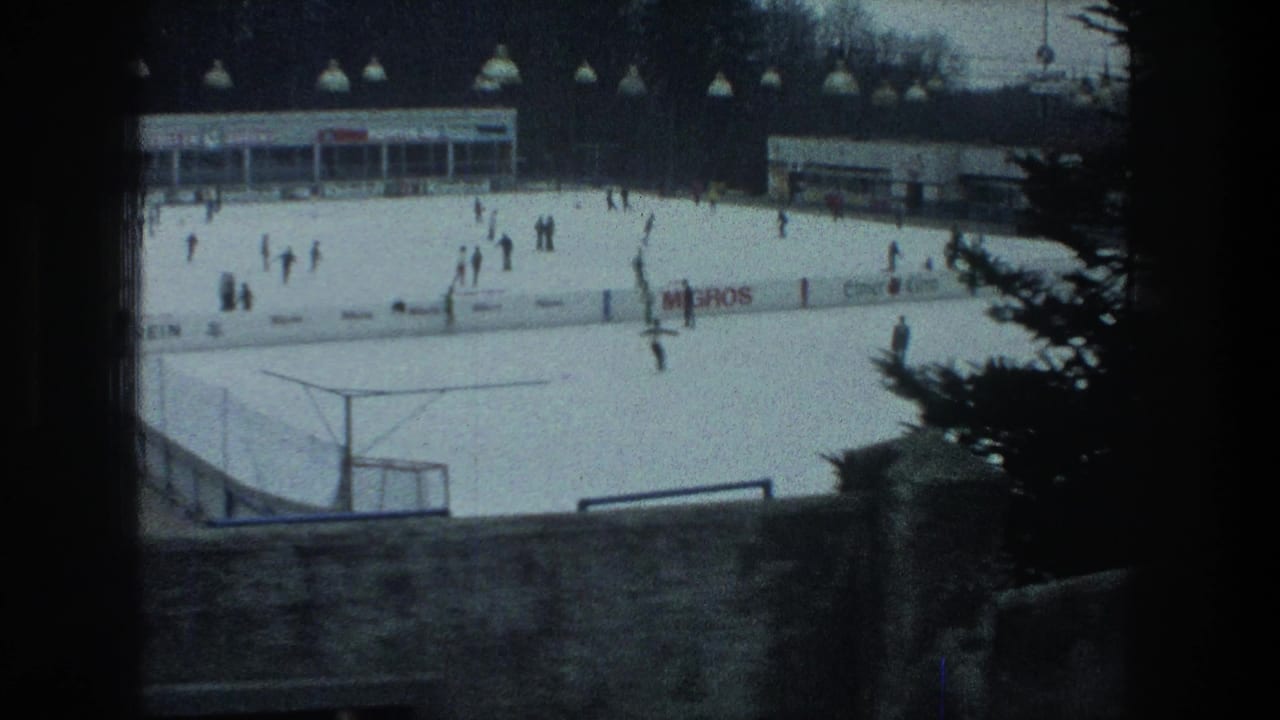 Wide 1976 Zurich home-movie view of skaters on an outdoor rink, with boards, signage, buildings, and a tree at frame edge.