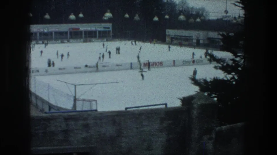 Wide 1976 Zurich home-movie view of skaters on an outdoor rink, with boards, signage, buildings, and a tree at frame edge.