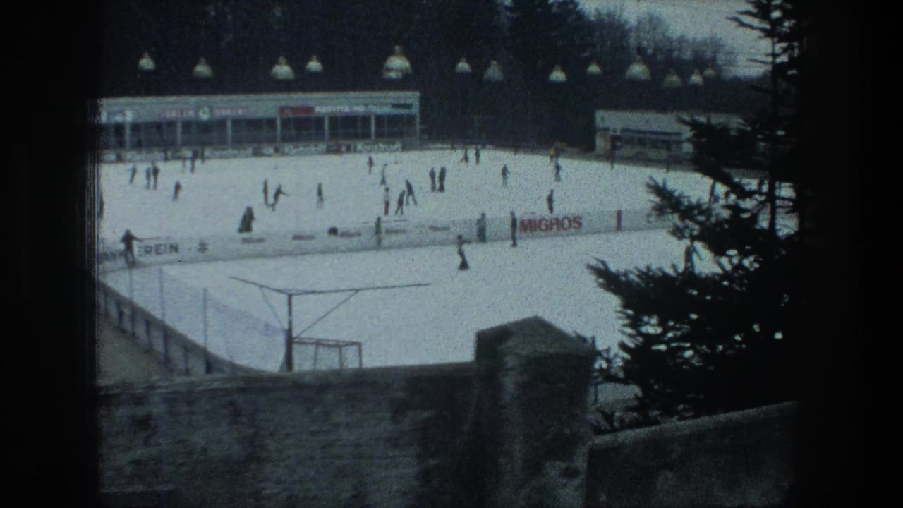 1976 Zurich outdoor ice rink seen from above, with skaters on the ice, hanging lights, a rink building, a stone wall, and pine branches.