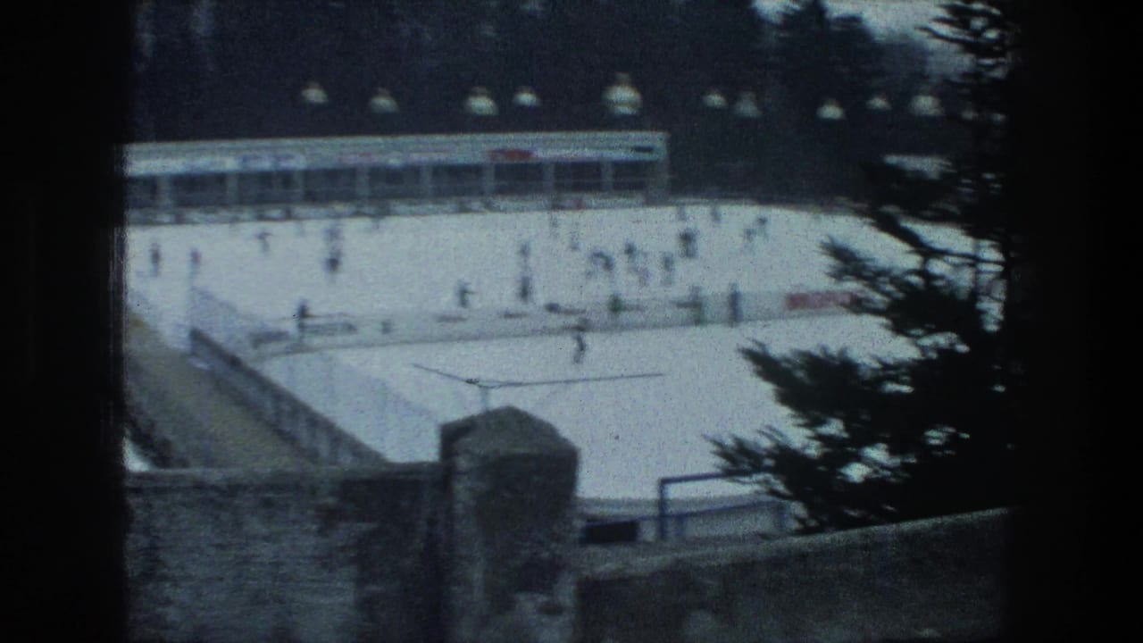 Soft 8mm view of a Zurich outdoor ice rink in 1976, with skaters, overhead lamps, a rink building, and dark pine branches in the foreground.
