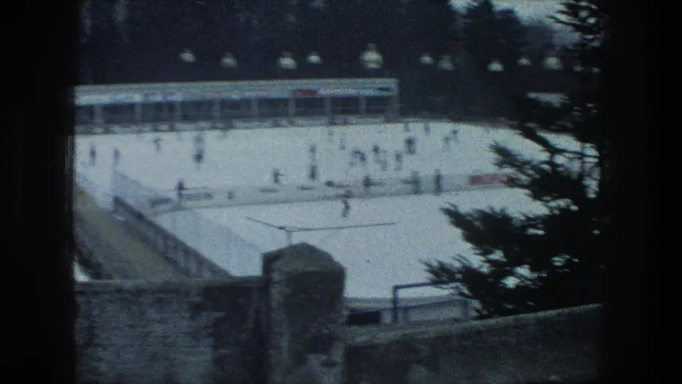 Soft 8mm view of a Zurich outdoor ice rink in 1976, with skaters, overhead lamps, a rink building, and dark pine branches in the foreground.