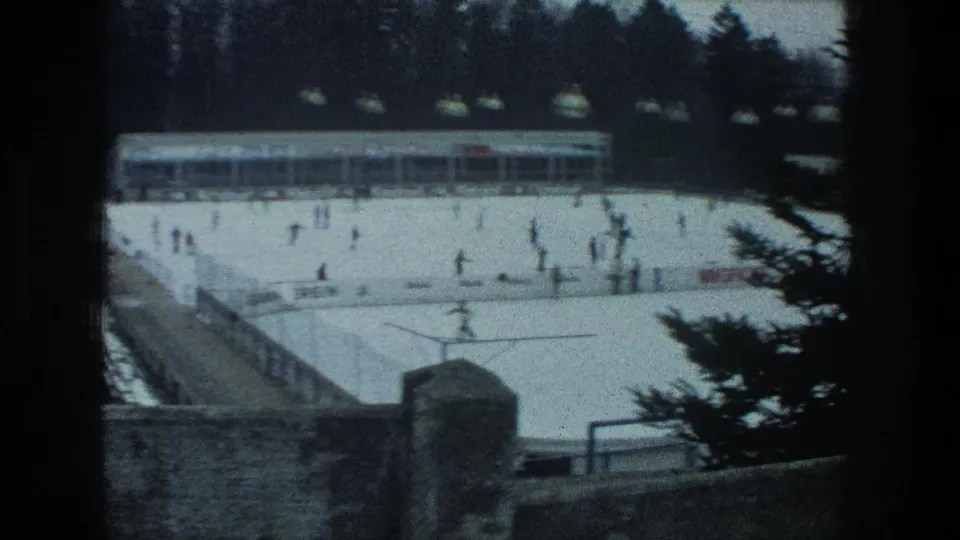 Elevated 1976 Zurich rink view with skaters, advertising boards along the barrier, hanging lights, a rink building, a stone wall, and pine branches.