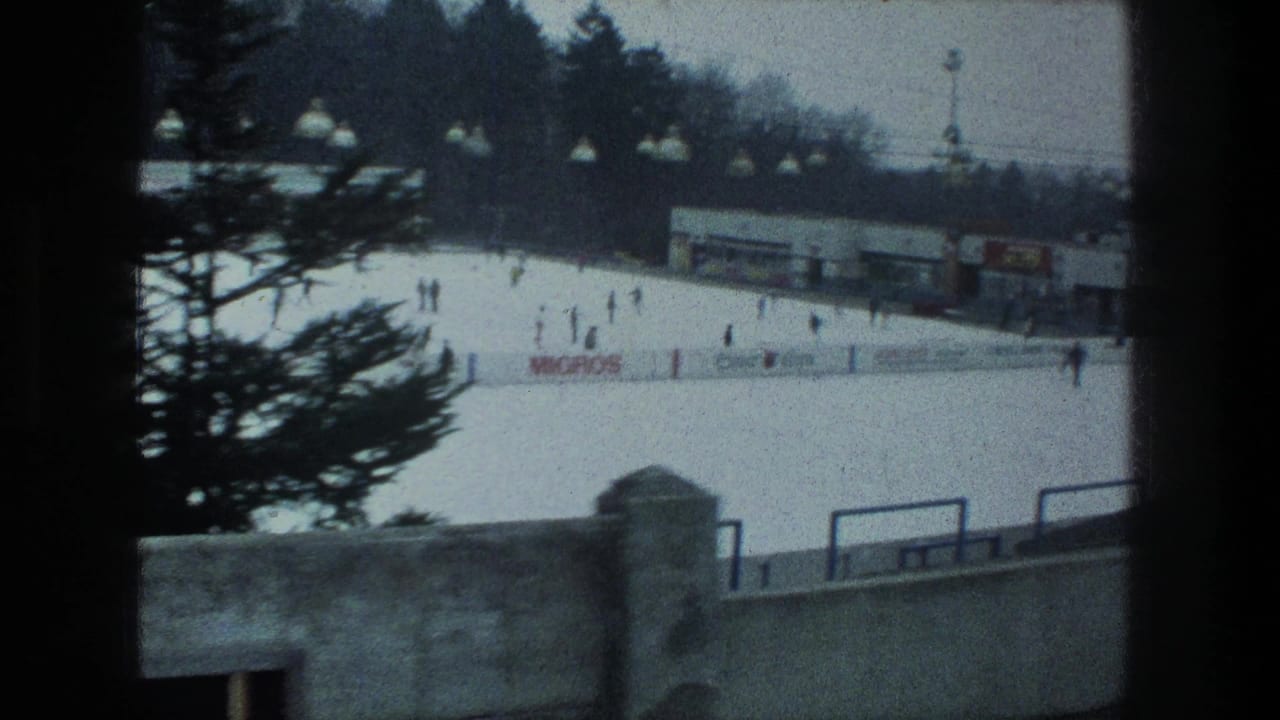Home-movie view of a Zurich outdoor rink in 1976, with skaters, advertising-style rink boards, a concrete wall and a tree crossing the frame.