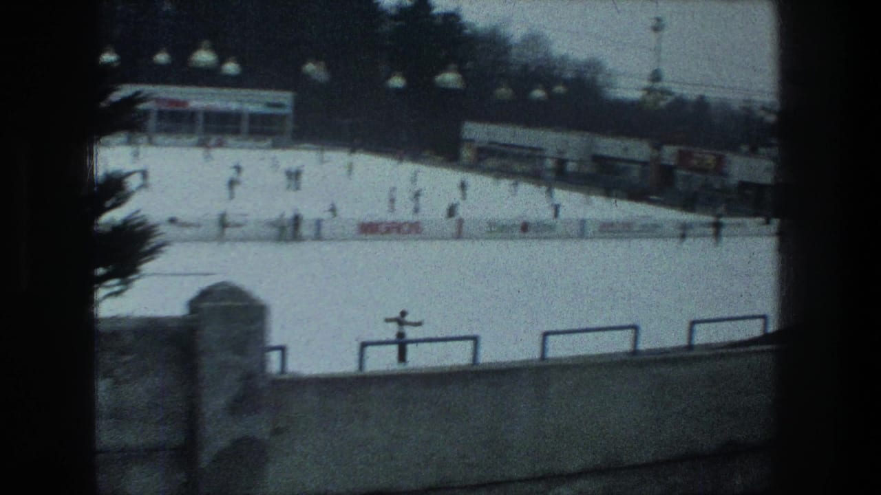 Wide 1976 Zurich outdoor skating rink view with distant figures on the ice, concrete foreground barrier and low buildings beyond.