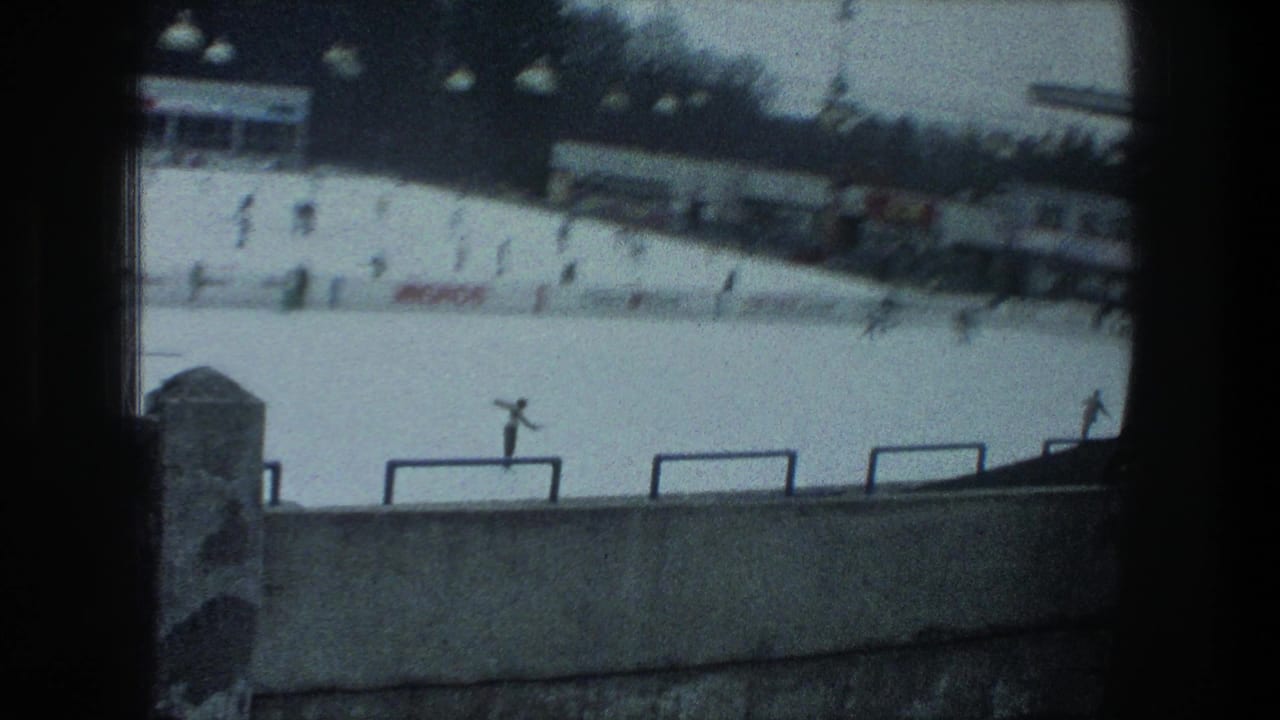 Outdoor ice rink with scattered skaters, barriers, buildings, and a Migros sign in the background.