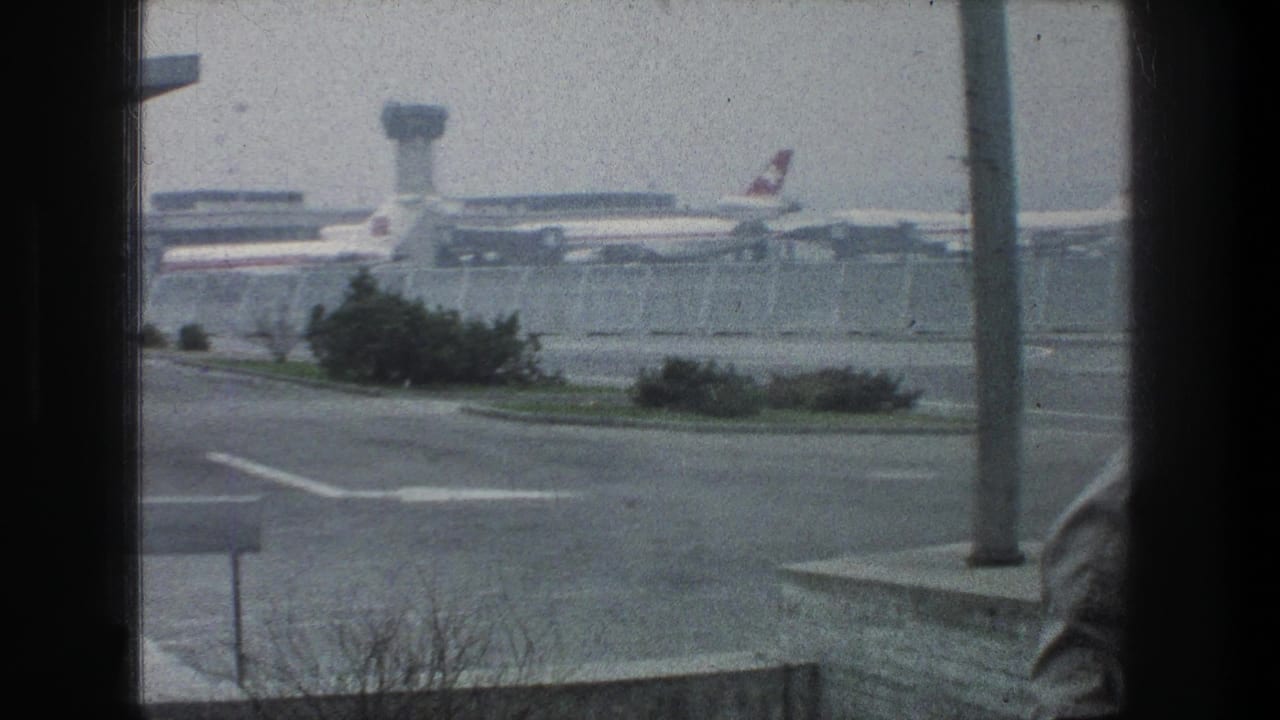 Home-movie frame from Zurich showing an airport-area scene with aircraft tails, control tower, roadway, shrubs, and a foreground pole.