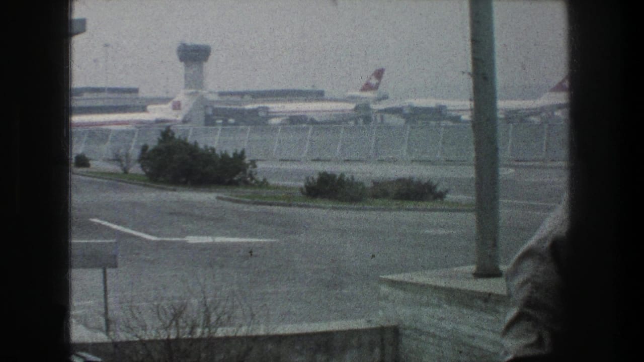 1976 Zurich Airport view through a window, with parked jets, control tower, fencing, roadway, shrubs, and a partial foreground figure.