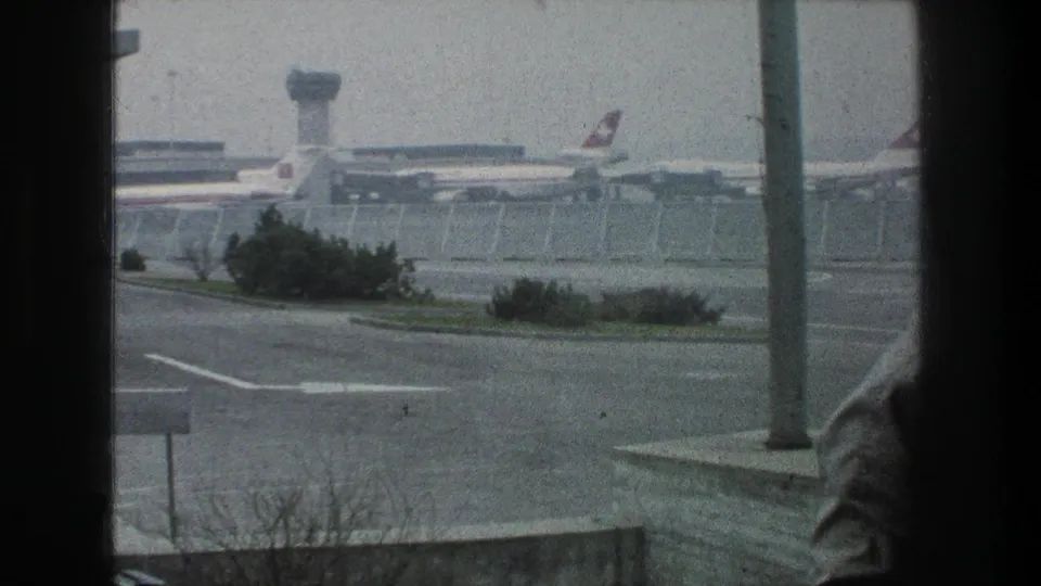 1976 Zurich Airport view through a window, with parked jets, control tower, fencing, roadway, shrubs, and a partial foreground figure.