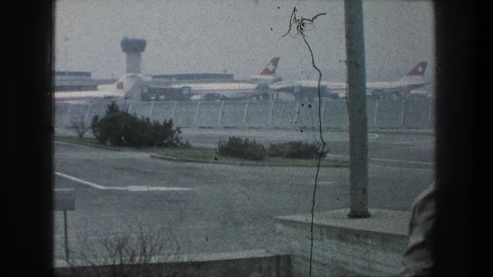 1976 Zurich Airport scene showing parked passenger jets and control tower beyond fencing, with a partial foreground figure near the window edge.