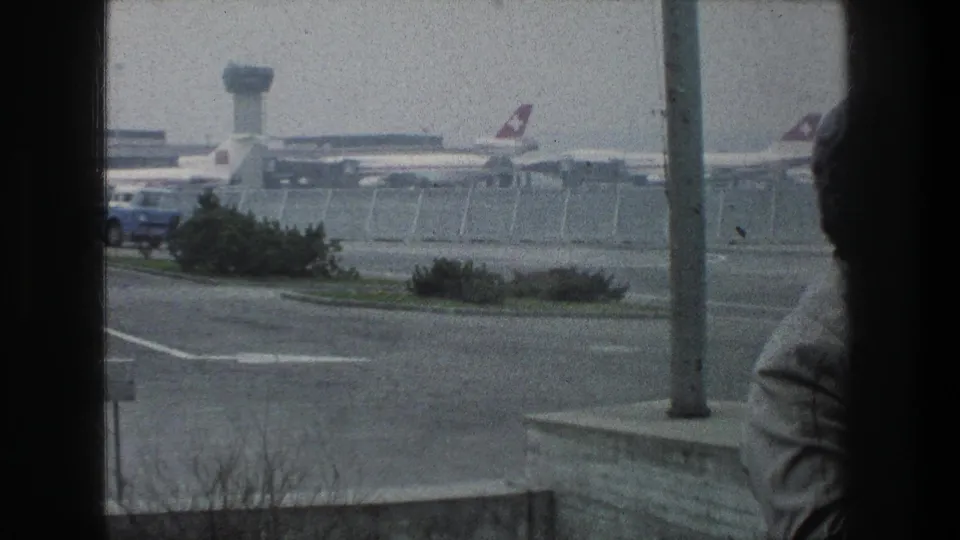 1976 Zurich Airport view with a blue and white shuttle bus crossing the foreground, parked jets behind fencing, and a partial figure at right.