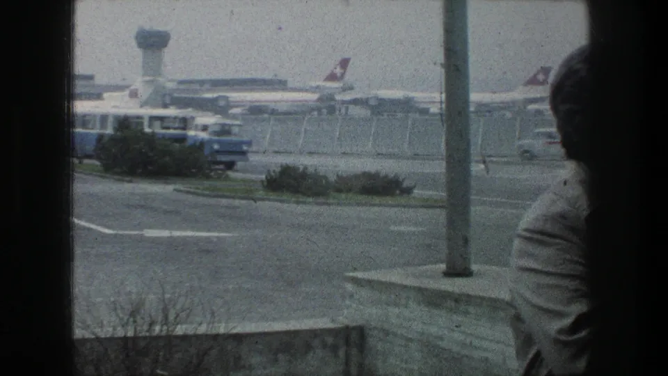 Late clip frame from 1976 Zurich Airport showing a shuttle bus, parked passenger jets, control tower, fencing, window frame, and partial foreground person.