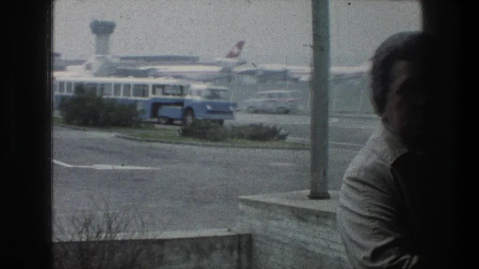1976 Zurich airport view through terminal glass showing a blue shuttle bus, parked Swissair-marked jets, and a partially visible observer.