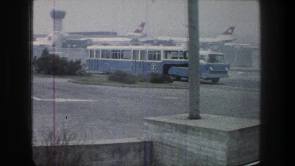 1976 Zurich airport tarmac view with a blue shuttle bus, parked Swissair-marked aircraft, terminal structures, and cloudy sky.