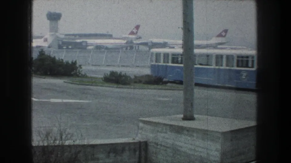Zurich airport in 1976 seen from behind terminal framing as a blue shuttle bus passes parked aircraft and ground structures.