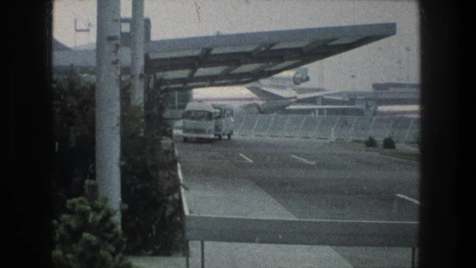 1976 Zurich airport view with a white bus on an access road, a modern canopy overhead, and jet aircraft visible past the fence.