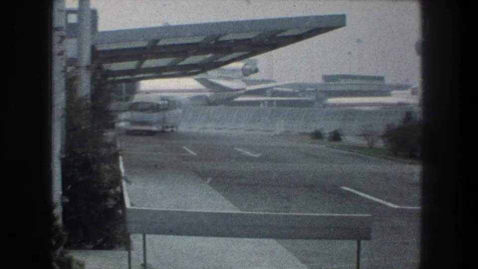 Zurich airport exterior in 1976, showing a bus near the left edge, roadway lanes, canopy structure, and aircraft at the terminal.