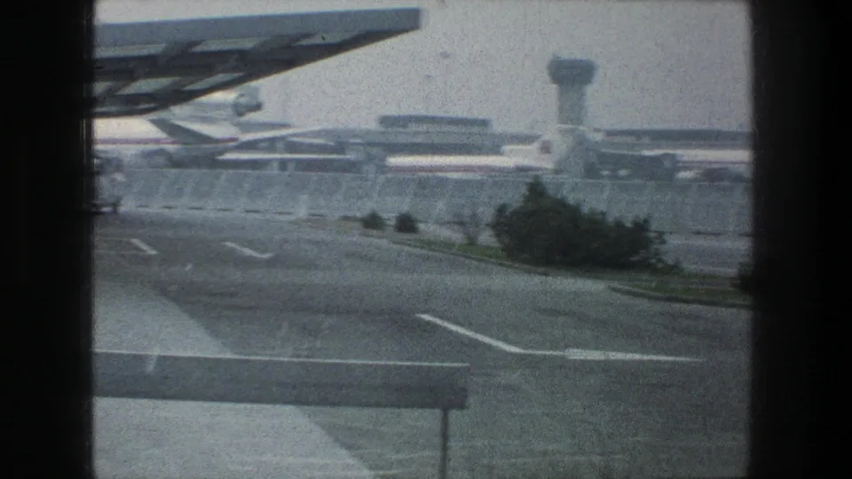 1976 Zurich airport scene with a curving roadway, fenced terminal area, parked aircraft, and a control tower under a hazy sky.