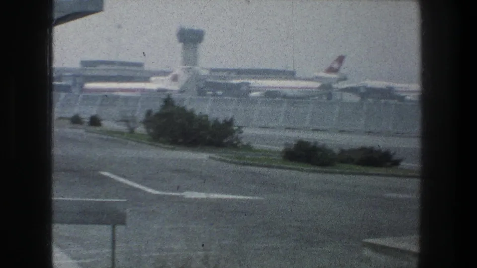 Zurich airport in 1976 with parked commercial aircraft, a visible control tower, fenced terminal buildings, and an empty foreground road.