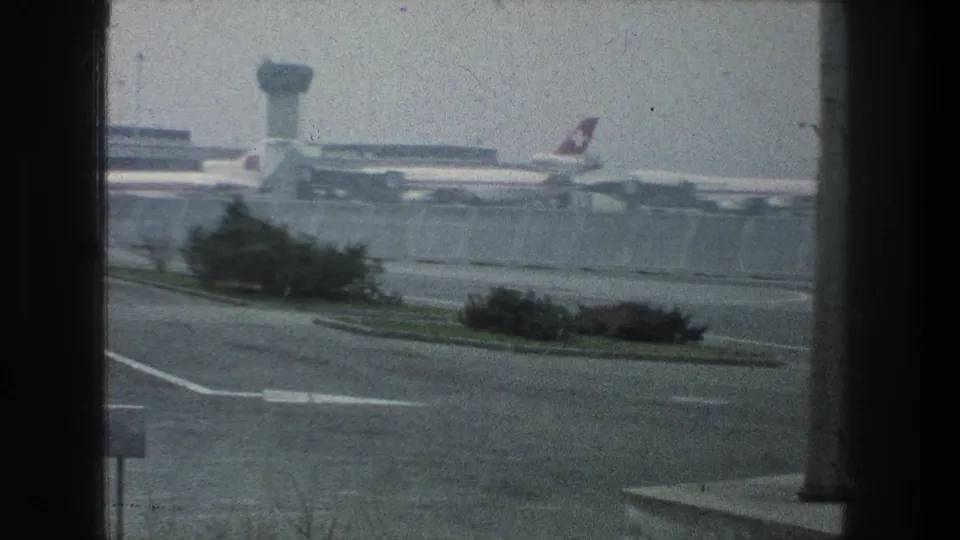 1976 Zurich Airport view through a window with Swissair aircraft tails, terminal structures, road pavement, shrubs, and a distant tower.