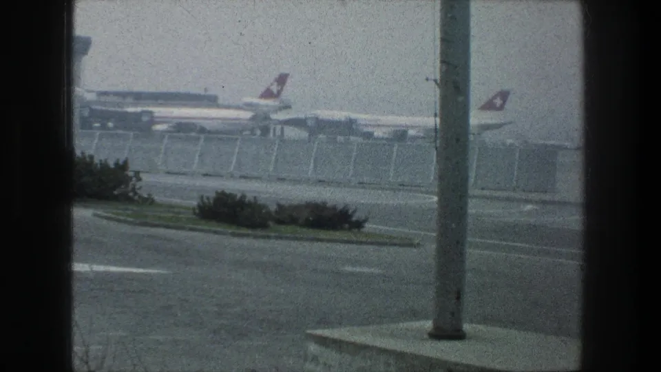 A Zurich Airport window-framed view shows Swissair aircraft by the terminal, with a light pole, road markings, shrubs, and hazy sky.