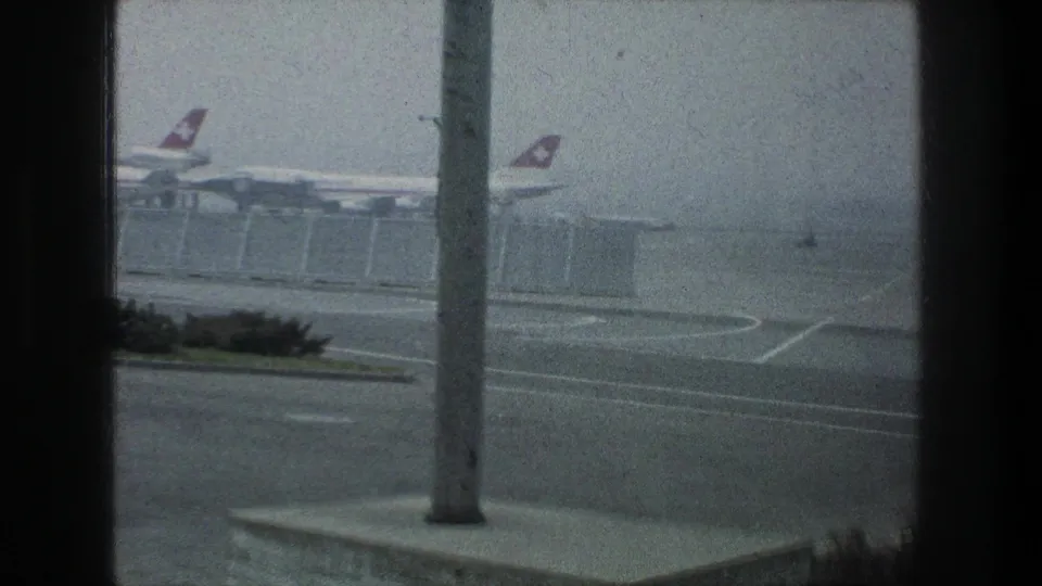 The 1976 Zurich Airport footage frames Swissair aircraft tails, a fenced terminal area, road curves, shrubs, and a foreground pole through glass.