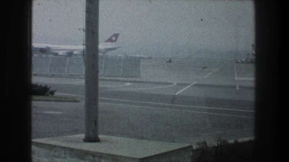 A hazy Zurich Airport frame shows a Swissair tail near the terminal, foreground pavement, a tall pole, dark window mullions, and roadside shrubs.
