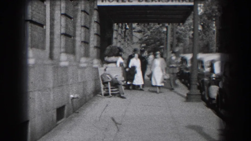 People in period clothing walk past the Hotel Berkshire entrance in Reading, Pennsylvania, with parked cars lining the curb.
