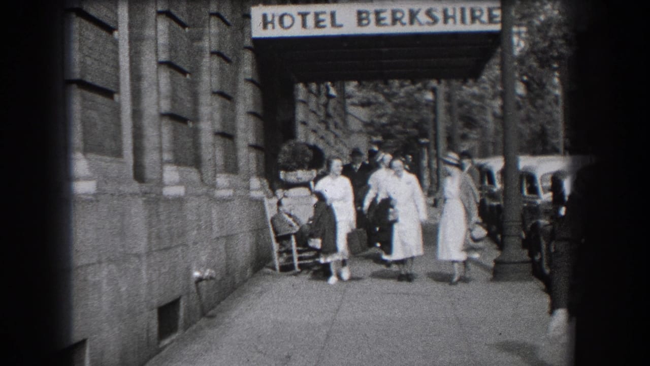 Home-movie frame of women walking along the Hotel Berkshire sidewalk as a seated man remains near the building wall.