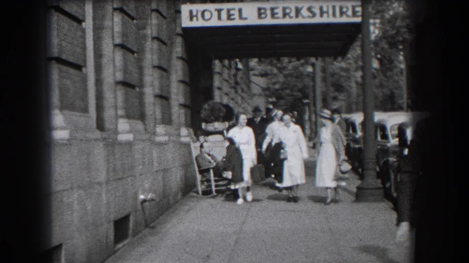 Home-movie frame of women walking along the Hotel Berkshire sidewalk as a seated man remains near the building wall.
