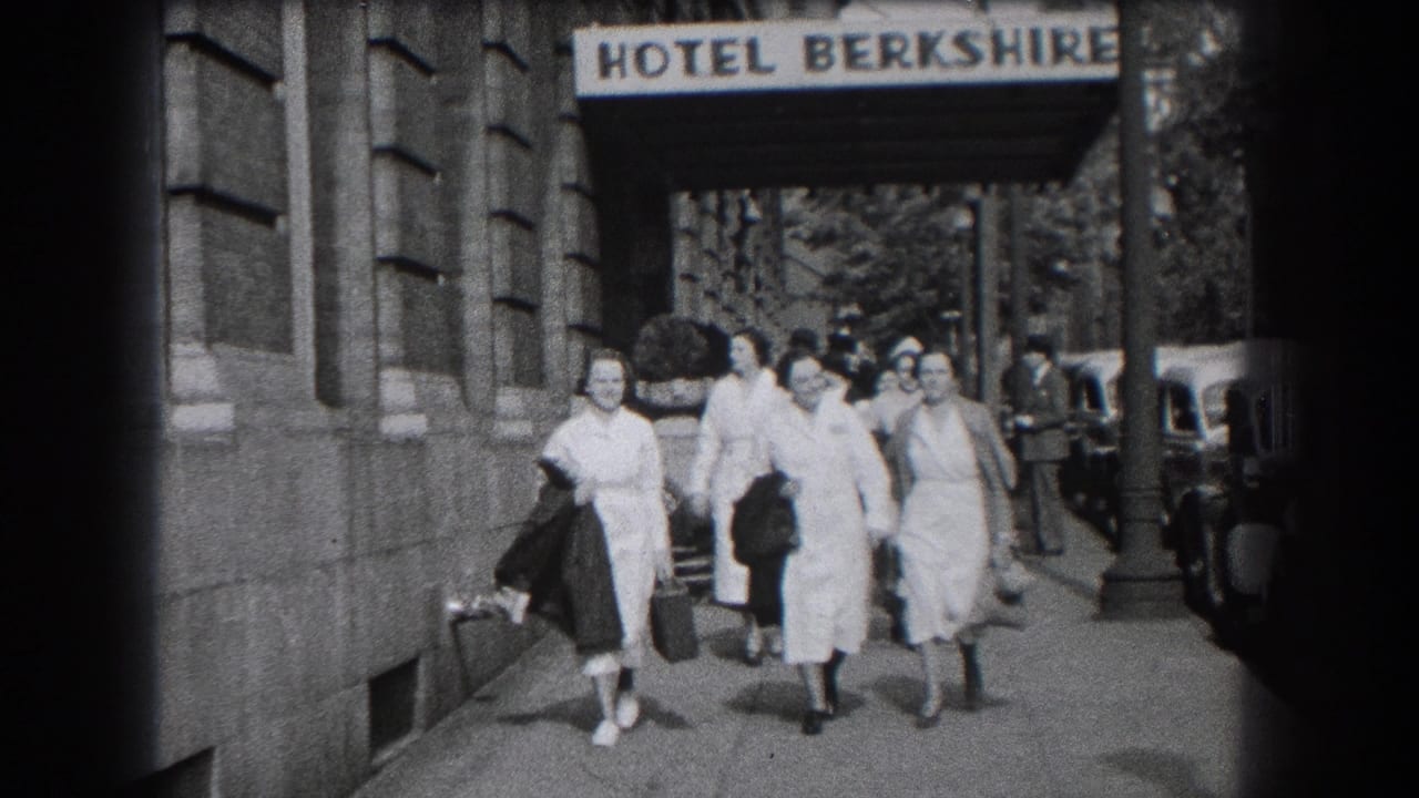 A group of women in white coats and dresses walk toward the camera near parked cars.