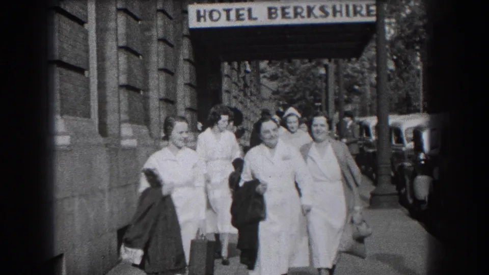 Close sidewalk frame of smiling women in nurse-style dress near the Hotel Berkshire sign, with vintage cars visible along the curb.