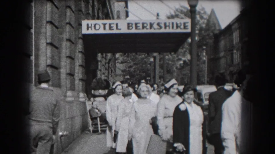 A group of nurses in white uniforms and caps walks near the Hotel Berkshire entrance, with sidewalk activity behind them.