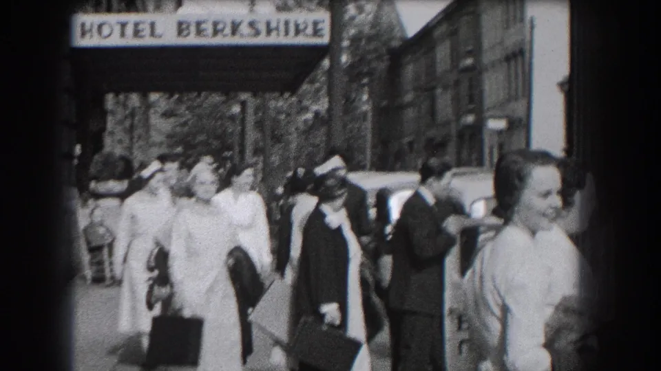 Nurses in white uniforms pass close to the camera outside the Hotel Berkshire, with pedestrians and parked cars in the background.