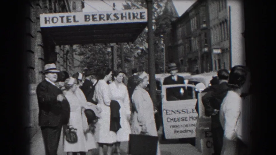 Adults in 1930s dress stand and walk near the Hotel Berkshire canopy, with a sidewalk signboard and vehicle in the background.