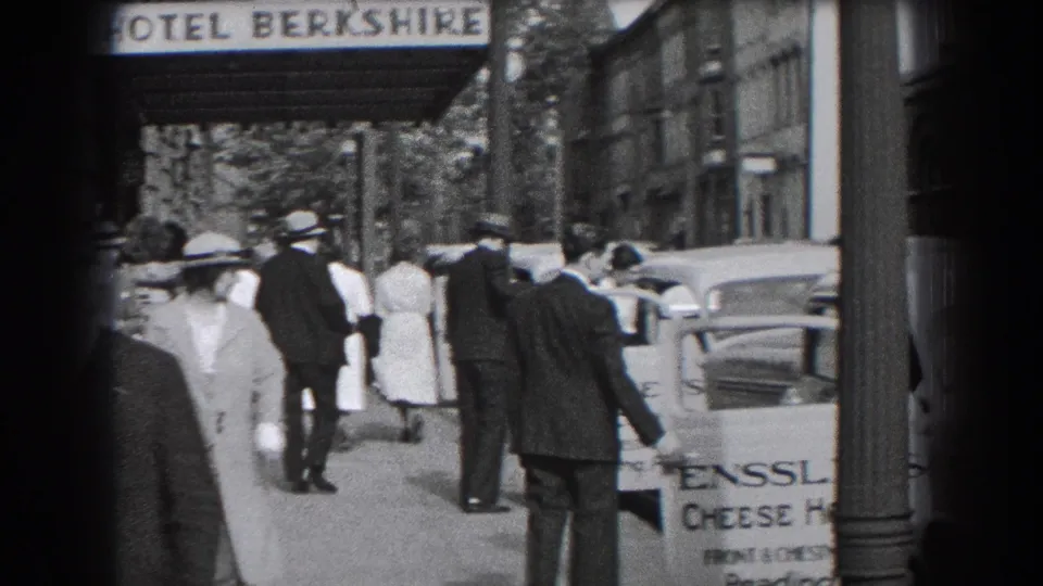 Pedestrians move down the sidewalk outside the Hotel Berkshire, with automobiles, building fronts, and a partial street signboard visible.