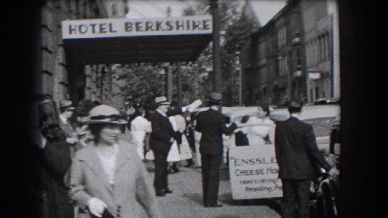 A woman in a hat walks toward camera near the Hotel Berkshire sign and parked vehicles.