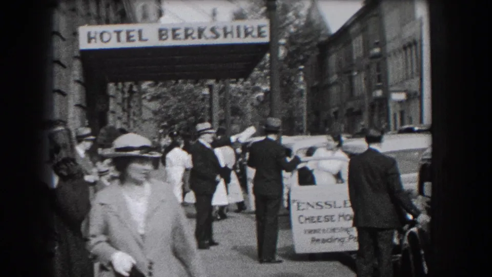Street-level 1938 Reading view with a woman approaching the camera, hotel signage overhead, pedestrians, and parked cars nearby.