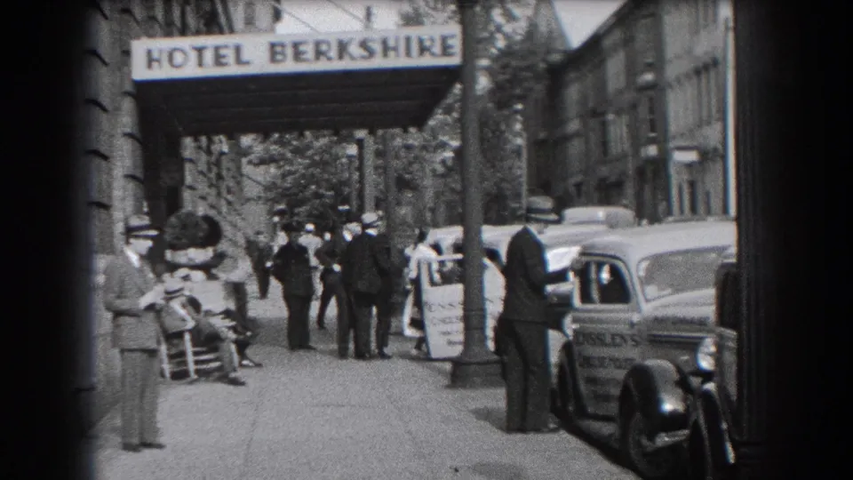 1938 home-movie frame showing men on the sidewalk outside Hotel Berkshire, with an automobile and Ensslen's Cheese House marking visible.