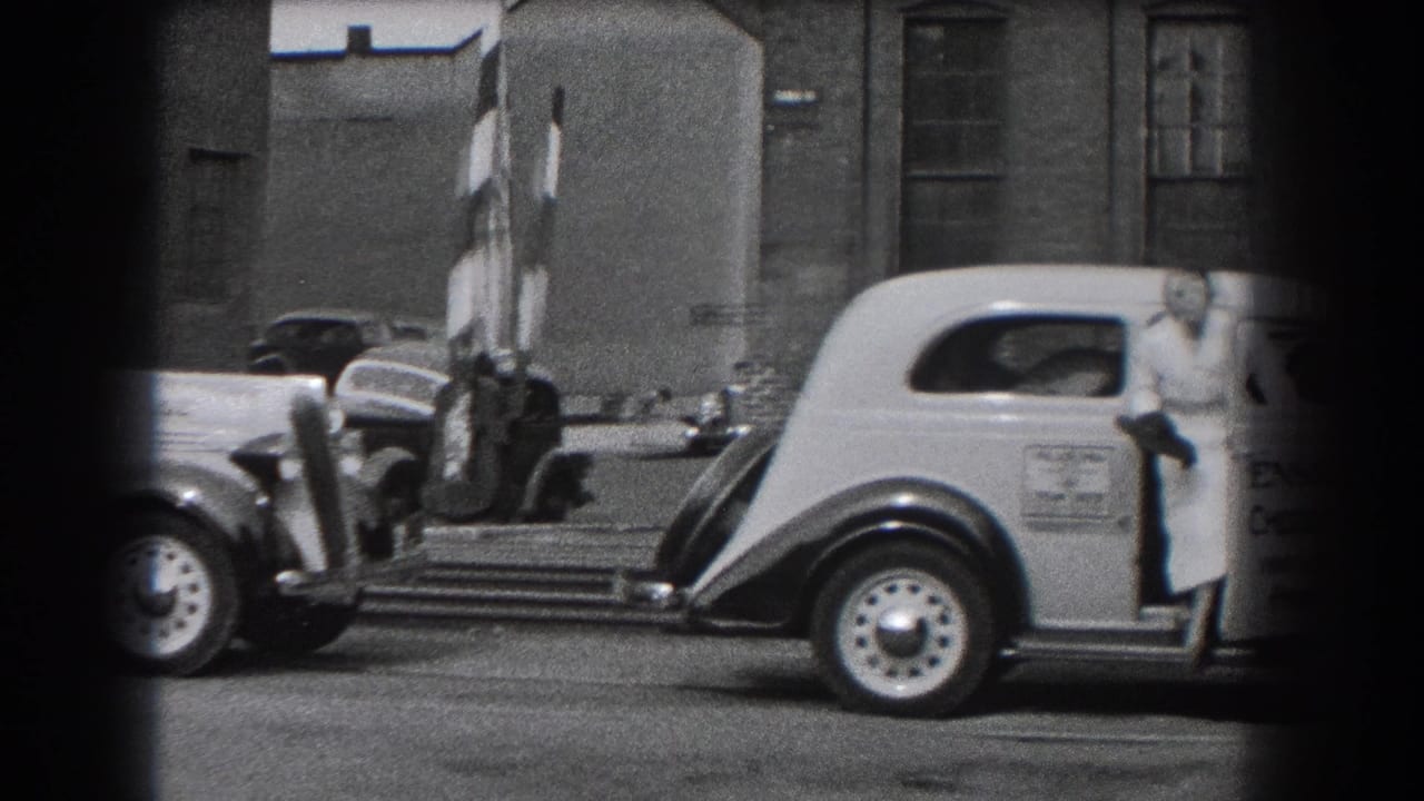Light-colored vintage car stopped by striped crossing barriers as a woman stands at the door.