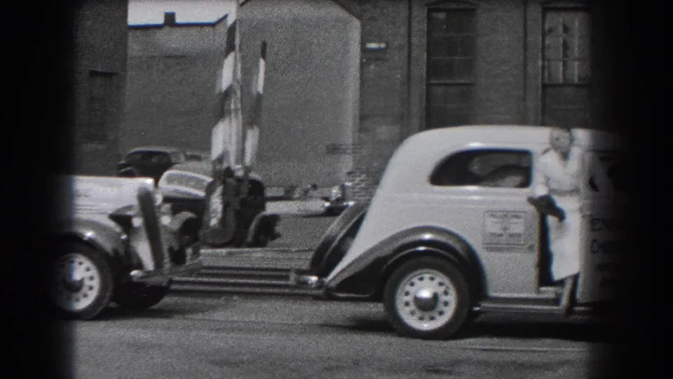 In 1938 Reading, Pennsylvania, a light-colored vehicle is stopped near striped crossing barriers while a woman in a long dress stands at the door.
