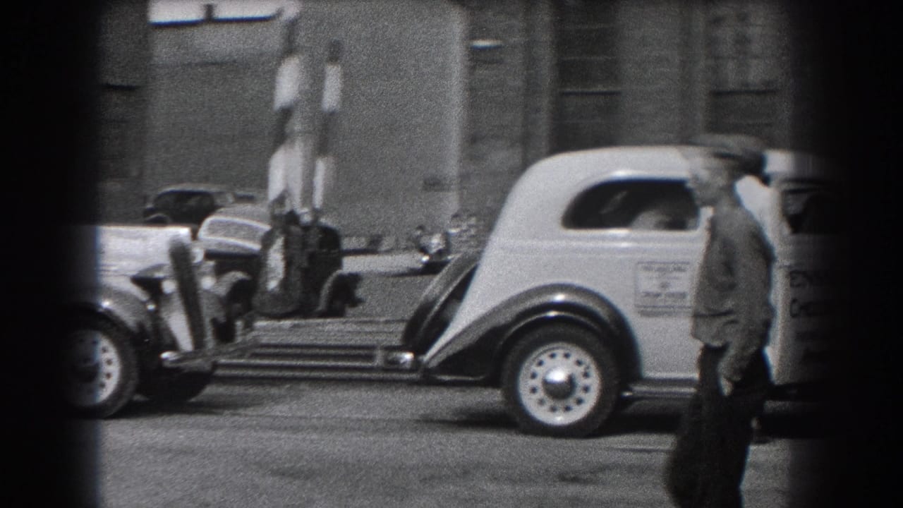 A man in a cap passes in front of a light-colored vehicle near striped barriers and parked cars in Reading, Pennsylvania.