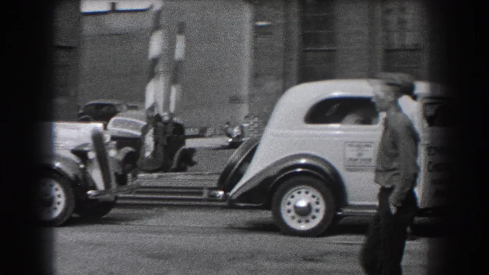 A man in a cap passes in front of a light-colored vehicle near striped barriers and parked cars in Reading, Pennsylvania.