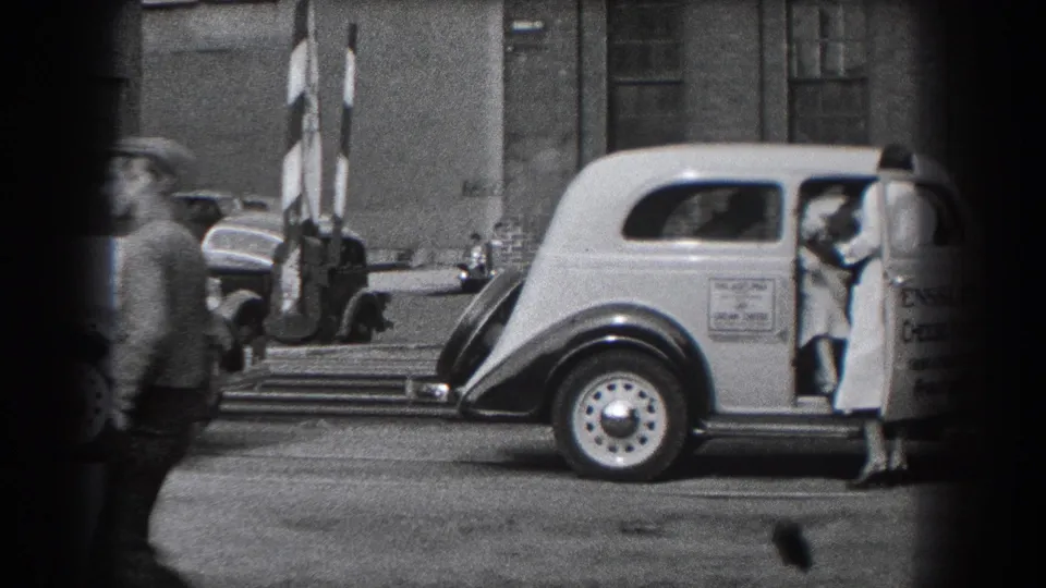 A woman in a long dress and heels stands beside the open door of the light-colored vehicle as a passerby moves across the edge of the frame.