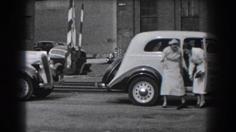 Two women in long dresses and heels stand beside the light-colored vehicle near striped crossing barriers in the Reading, Pennsylvania street scene.