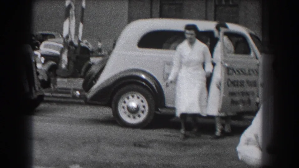 In Reading, Pennsylvania, women in white uniforms move beside a 1930s Ensslen's Cheese House delivery car on a city street.