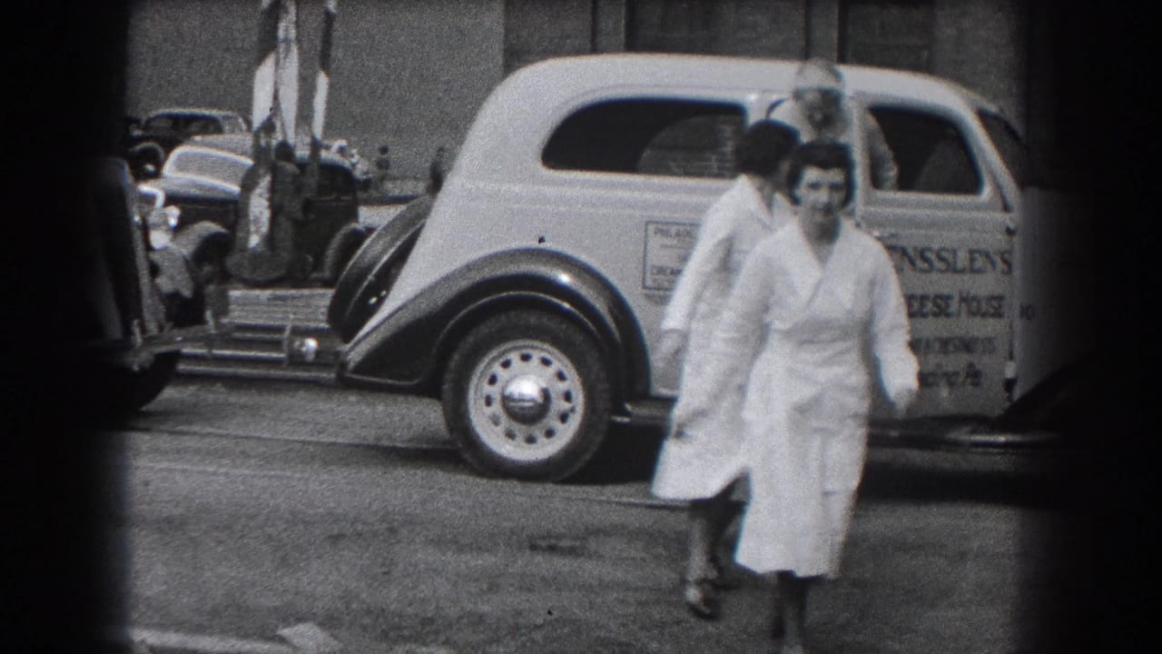 A woman in a white uniform moves past a vintage car with business lettering.