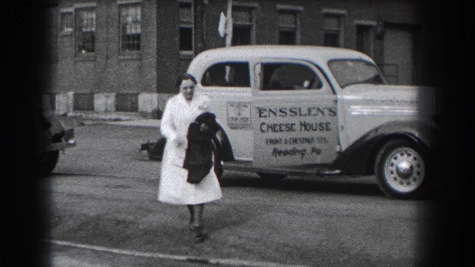 In Reading, Pennsylvania, a woman in a white dress walks near a parked Ensslen's Cheese House delivery car on a city street.